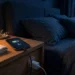 A smartphone plugged into a charger on a bedside table at night, its screen showing a battery icon and a small moon, in a dark bedroom lit by a warm lamp