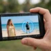 A hand holding a smartphone showing a beach photo being edited, with a glowing selection ring around a stranger in the background about to be erased.
