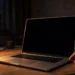 A laptop with a dark screen on a desk at night, a hand reaching toward the power button