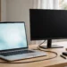 A laptop with a lit screen on a wooden desk, connected by a cable to a second external monitor that remains completely black