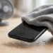 A wet smartphone tilted so its charging port faces down, water dripping away, being patted dry with a grey microfiber cloth, with a desk fan in the background