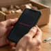 Hands holding a smartphone showing the "Erase All Content" option, with a removed SIM card, SIM ejector tool, and shipping box on a wooden desk
